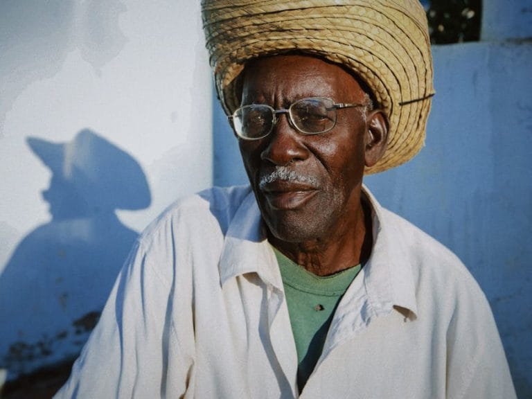 Vieil homme au chapeau de paille, devant un mur ensoleillé, avec une ombre marquée.